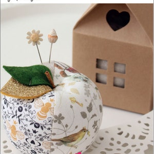 May include: A white and gold apple-shaped pincushion with a green felt leaf on top. The pincushion is decorated with a floral pattern and a small bird. The pincushion is sitting on a white doily. The text "APPLE PINCUSHION" is at the top of the image, and the text "by Nic Vaughan" is below it. The text "Craftapalooza Designs" is at the bottom of the image.