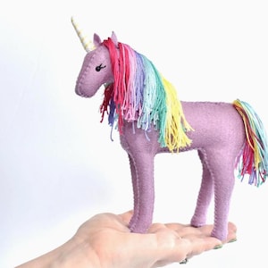 May include: A purple felt unicorn with a rainbow mane and tail. The unicorn has a gold horn with white stripes.