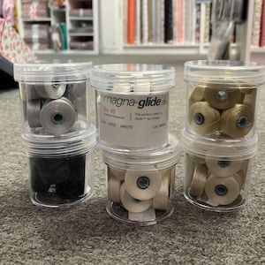 May include: Six clear plastic containers filled with spools of white, gray, and beige thread. The containers are labeled "magna-glide" and "No. 40 Tibal Polyester".