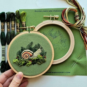 May include: An embroidery kit featuring a snail design. The kit includes a green fabric hoop with a snail and foliage embroidery, embroidery floss in various shades of green, and the text "GUIDELINES ARE PERMANENT".