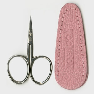 May include: A pair of small, silver-colored scissors with curved handles and pointed blades, next to a pink leather case. The case is oval-shaped and has a stitched border.