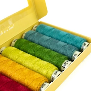 May include: A yellow box containing spools of colorful thread. The spools are in shades of red, yellow, green, and blue. The box has the text "One Stitch at a Time!" printed on the inside.