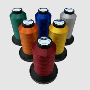 Op de afbeelding: Zes spoelen Glide 60 garen in verschillende kleuren: groen, oranje, blauw, geel, zilver en rood. Elke spoel heeft een zwart label met witte tekst waarop "Glide 60" staat.
