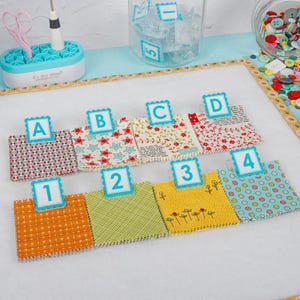 May include: A craft project setup with fabric swatches labeled A, B, C, D, and 1, 2, 3, 4. The swatches display various patterns and colors, including red, blue, yellow, and green. Crafting tools and supplies are visible in the background.