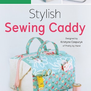 May include: A turquoise and white sewing caddy with floral print and a pink zipper. The caddy has two handles and a pocket with a measuring tape, scissors, and a note. The caddy is sitting on a white surface with a pink and white pincushion and a spool of thread.