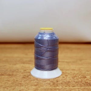 Könnte beinhalten: Eine Spule blau-grauen Fadens mit gelbem Oberteil und weißem Sockel. Der Faden ist eng um die Spule gewickelt und bildet eine zylindrische Form. Die Spule steht auf einer Holzoberfläche.