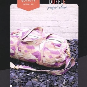 May include: A pink and white camouflage print duffel bag with a gold zipper and a brown leather strap. The bag is lying on a bed of small, dark grey stones. The text "REIGN DUFFLE project sheet" is printed on the top of the image.