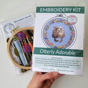 May include: Embroidery kit with a beginner's guide, a wooden hoop, embroidery floss, needles, and a printed design of two otters holding hands in a floral wreath. The kit includes instructions and an online video tutorial. "Otterly Adorable" is the title of the design.