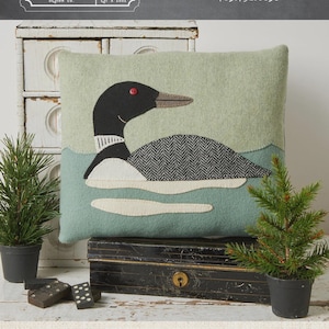 May include: A decorative cushion with a loon design. The cushion has a light green background with a loon illustration in black, white, and grey. The cushion measures 28 cm by 33 cm.