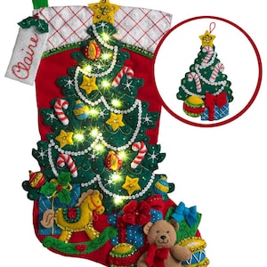Pode incluir: Uma meia de Natal vermelha com um design de árvore de Natal em feltro, adornada com bengalas doces, estrelas e luzes. Inclui um cavalo de baloiço, um urso de peluche e presentes. Um pequeno enfeite de árvore é mostrado num círculo. O nome "Claire" é bordado.