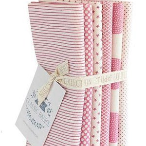 Peut inclure: Un ensemble de coupons de tissu aux motifs variés rose et crème, dont des rayures, des pois et des étoiles. Les tissus sont liés par un ruban avec l'inscription "Tilde Quilts Collection". Une étiquette "Classic Basics" est attachée avec un nœud.