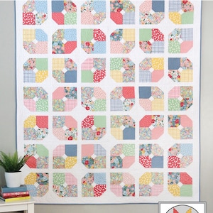 May include: A quilt pattern called "Make Believe" featuring a patchwork design with various floral and geometric prints in pink, blue, green, yellow, and white. The quilt is hanging on a white wall with a white table and a plant in a white pot in the foreground. The quilt is made by "a bright corner".
