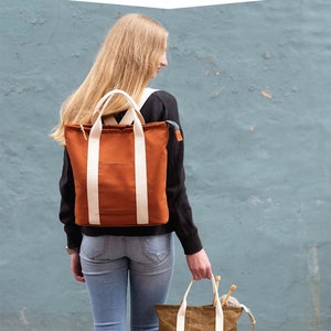 Może przedstawiać: Kobieta nosi brązowy plecak z białymi paskami i trzyma brązową torbę z białymi paskami. Torba ma zamek błyskawiczny i jest wypełniona drutami do robótek na drutach. Tekst "BUCKTHORN backpack & tote" znajduje się w górnej części obrazu. Tekst "noodlehead" znajduje się w dolnej części obrazu.