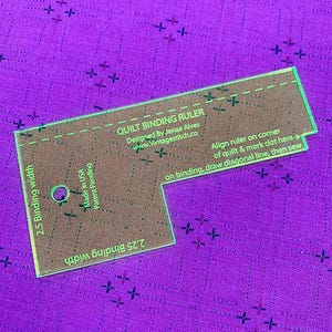 May include: A clear acrylic quilt binding ruler with green edges, resting on a purple fabric. The ruler has text that reads "QUILT BINDING RULER" and "2.5 Binding width". It also includes instructions for use and is marked "Made in USA".
