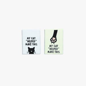 May include: Two rectangular fabric labels. One is white with a black cat silhouette and the text "MY CAT 'HELPED' MAKE THIS". The other is light green with a black cat paw and the same text.