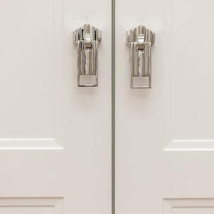 May include: Two silver cabinet door pulls with a geometric design. The pulls are mounted on a white cabinet door.
