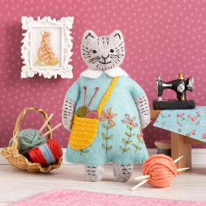 May include: A grey felt cat doll wearing a light blue dress with embroidered flowers and a yellow bag containing yarn and knitting needles. The cat has a white collar and a smiling face.