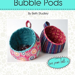May include: Two fabric hanging baskets, one teal and one pink, with a starburst pattern. The baskets are 5 1/2 inches wide, 5 inches high, and 5 inches deep. The baskets are labeled "Bubble Pods" and are made by Beth Studley. The text "love from beth" is written in a cursive font on a teal oval.