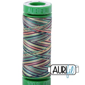 Pode incluir: Uma bobina de fio Aurifil 100% algodão em um padrão multicolorido variegado com tons de azul-esverdeado, roxo, dourado e marrom. A bobina é verde com o logotipo Aurifil e o texto "100% Algodão" impresso nela.
