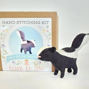 May include: A handmade felt skunk stands in front of a craft kit box. The skunk is black and white with a pink nose. The box reads "Hand Stitching Kit" and "Felt Skunk Sewing" with the words "Learn to Sew" at the bottom.