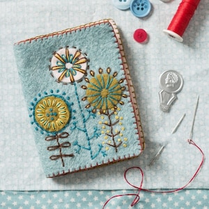 May include: A blue felt needle case with a stitched design of three flowers in yellow, green, and blue. The case has a brown stitched border.