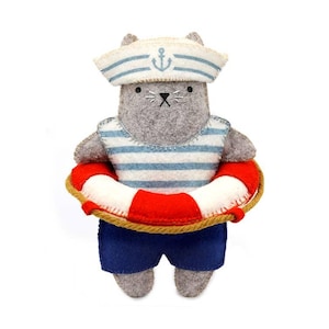 May include: A felt cat plush toy wearing a white and blue striped sailor shirt, a matching sailor hat with a blue anchor emblem, and dark blue shorts. The cat is positioned inside a red and white life preserver ring with a brown rope detail.