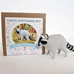 May include: A gray felt raccoon toy stands next to a cardboard box labeled "HAND STITCHING KIT". The box features a raccoon image and the text "RACCOON SEWING KIT" and "Learn to Sew". The raccoon toy has a black mask and striped tail.