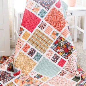 May include: A quilt with a geometric pattern in shades of red, yellow, blue, and white. The quilt is made of various floral fabrics. The quilt is folded and lying on a light brown surface. The text "Craftsman" is in a blue banner at the top of the image. The text "Amy Smart - Diary of a Quilter" is in a white banner at the bottom of the image.