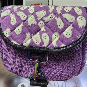 Op de afbeelding: Een kleine, paarse crossbodytas met een gewatteerde flap met witte spookontwerpen. De tas heeft een gebreide body, een zwarte rand en een regenboogkleurige metalen sluiting. De tas ligt op een naaimachine.