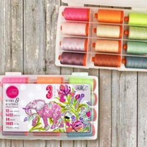 Op de afbeelding: Een set van 12 spoelen borduurgaren in een transparante kunststof opbergdoos. Het garen is verkrijgbaar in verschillende kleuren, waaronder roze, oranje, geel, groen en blauw. De doos heeft een label met de tekst "Threads & Trifles" en toont een roze olifantmotief.