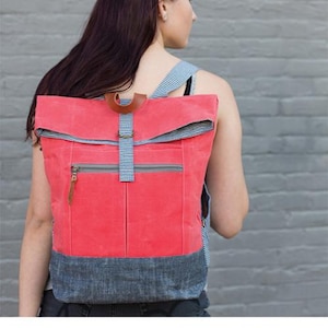 Może przedstawiać: Jasnokoralowy plecak z klapą na górze i paskiem w szachownicę w kolorze szarym i białym. Plecak ma kieszeń z zamkiem błyskawicznym z przodu i szary dolny panel. Tekst "RANGE BACKPACK" i "noodlehead" jest wydrukowany pod obrazem.