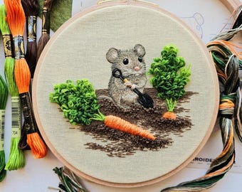 May include: A brown mouse with a small shovel embroidered on a white fabric in a wooden embroidery hoop. The mouse is surrounded by green plants and orange carrots.