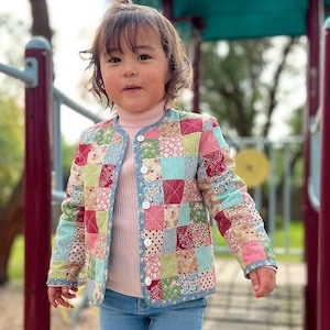 Może przedstawiać: Młoda osoba w kolorowej kurtce w patchworku, różowym golfie i niebieskich dżinsach. Kurtka ma różne wzory kwiatowe i geometryczne w odcieniach różowego, czerwonego, niebieskiego, zielonego i białego.