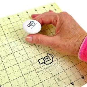 以下が含まれることがあります： 白い円形のキルトツールで、上部に黒い文字「QS」があります。このツールは、黄色と白のグリッドパターンが印刷されたキルト定規で使用されています。定規には「QuiltersSelect.com」というテキストが印刷されています。