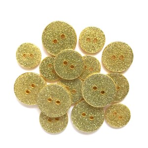 May include: A collection of gold glitter buttons in various sizes. Each button is round with two small holes for sewing. The buttons have a sparkly, textured surface and are scattered on a white background.