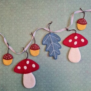 May include: A felt garland with autumnal shapes. The garland features red mushrooms with white spots, blue leaves, and acorns with brown tops and yellow bottoms. The shapes are strung on a ribbon against a patterned background.