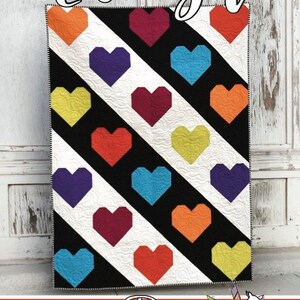 Könnte beinhalten: Ein Quiltmuster mit den Worten "luv ya" oben. Der Quilt besteht aus schwarzen, weißen und bunten herzförmigen Stoffstücken. Der Quilt wird von Running Doe Quilts für Villa Rosa Designs hergestellt.