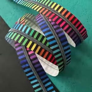 Könnte beinhalten: Ein farbenfroher Reißverschluss mit Regenbogenstreifen. Das Reißverschlussband weist horizontale Streifen in verschiedenen Farben auf. Die Metallzähne sind silberfarben. Der Reißverschluss ist aufgerollt und liegt auf einer türkisfarbenen und schwarzen Oberfläche.