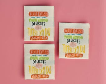 May include: Three rectangular fabric labels with care instructions for quilts. The labels are white with colorful text that reads "QUILT CARE INSTRUCTIONS MACHINE WASH COLD DELICATE TUMBLE DRY LOW SNUGGLE OFTEN."