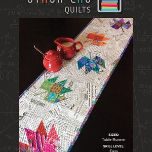 May include: A colorful quilt pattern called "Pressed Leaves" by Stash Lab Quilts. The quilt is a table runner with a design of colorful leaves on a white background. The quilt is shown on a black surface with a red creamer and sugar bowl.