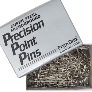 May include: A box of Prym Dritz Corporation Super Steel Microground Precision Point Pins. The box is silver with black text and the pins are silver.