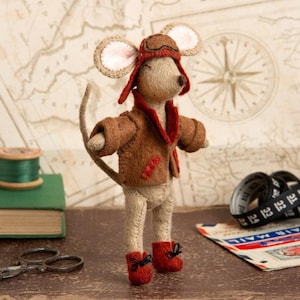 May include: A felt mouse wearing a brown aviator jacket with red accents, a brown leather aviator hat with a brown visor and goggles, and red boots. The mouse is standing on a brown surface.