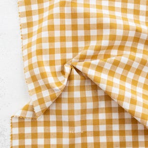 May include: Close-up of a folded fabric with a yellow and white gingham pattern. The fabric is a medium weight and has a soft texture. The pattern is a classic, timeless design, perfect for home decor or apparel projects.