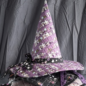 May include: A stack of four purple, black, and pink quilted witch hats with a black ribbon and a potion print. The hats are on a gray background.