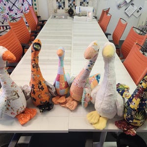 Op de afbeelding: Een verzameling handgemaakte ganzenknuffels in diverse kleurrijke stoffen. De ganzen hebben oranje snavels en voeten, met patchwork, bloemen en andere patronen. Ze zijn gerangschikt op een witte tafel.