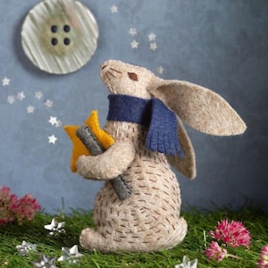 May include: A brown felt bunny wearing a blue scarf holds two yellow felt stars. The bunny is looking up at a white button with three black buttons on it, which is against a blue background with silver glitter stars.