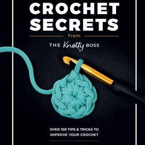 May include: A book cover with the title "Crochet Secrets from The Knotty Boss" and a teal crochet flower with a crochet hook. The book cover is black with white text and a white border. The text "Over 100 tips & tricks to improve your crochet" is below the title. The author's name, "Anna Leyzina", is at the bottom of the cover.