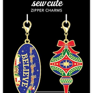 May include: Two gold zipper charms with a Christmas theme. One charm is a blue oval with the word "BELIEVE" and a scene of Santa's sleigh flying through the night sky. The other charm is a red, green, and gold ornament with a star in the center.