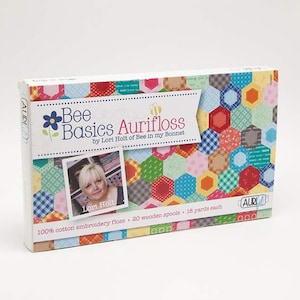 Op de afbeelding: Een doos Bee Basics Aurifloss borduurgaren van Lori Holt van Bee in my Bonnet. De doos heeft een kleurrijk patchwork ontwerp met zeshoeken en een foto van Lori Holt. De tekst op de doos luidt "Bee Basics Aurifloss by Lori Holt of Bee in my Bonnet" en "100% katoenen borduurgaren - 20 houten spoelen - 18 yards per spoel".