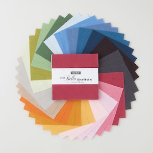 Op de afbeelding: Een waaier van 24 stoffen vierkanten in verschillende kleuren, waaronder groen, blauw, bruin, rood, oranje, geel en roze. De vierkanten zijn in een cirkel gerangschikt met een groter rood vierkant in het midden. Op het rode vierkant staat de tekst "Cozy Bella Solids by Moda".